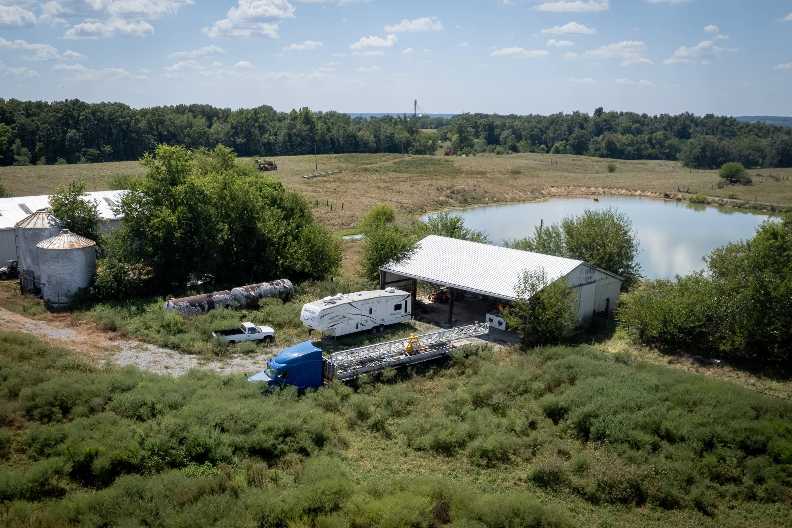 A truck with a large machine attached to it sits in a field in a rural setting, next to a camper van, a couple of buildings, silos and a pond.