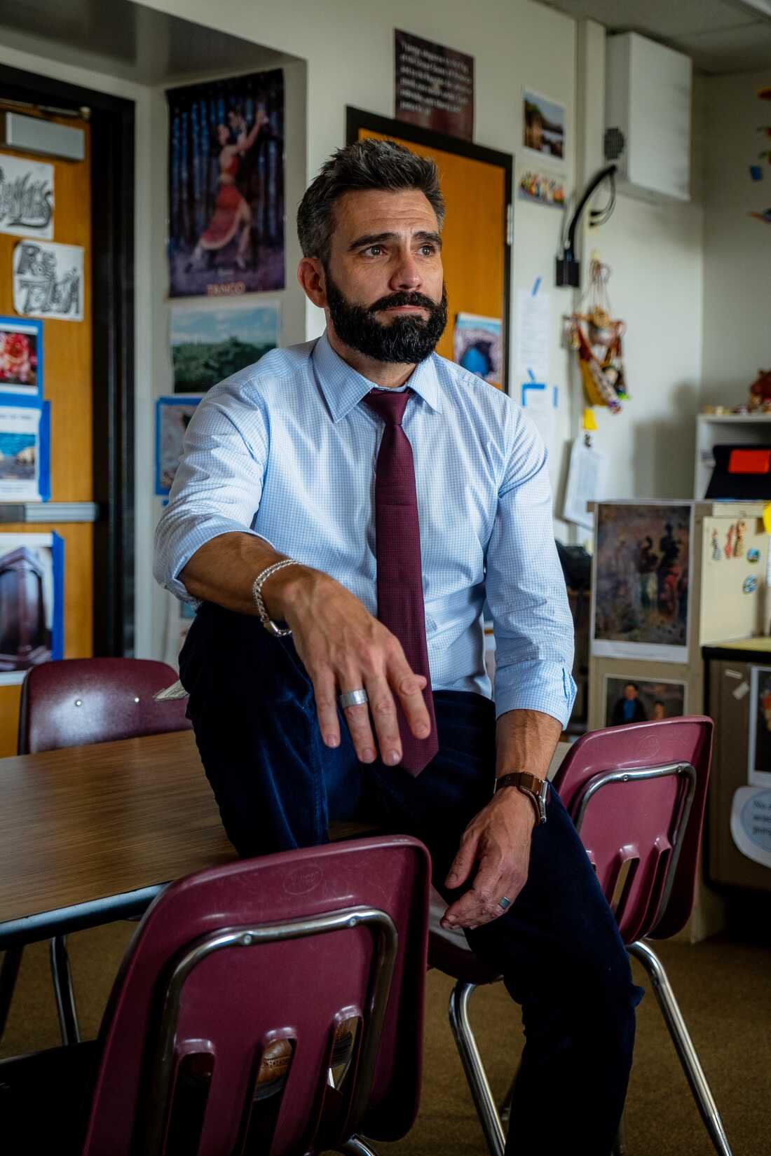 Jesse Kwakenat sits on a desk in his classroom. He has a beard and is wearing a button-down shirt and a tie. The classroom has posters on the walls and on the back of the door.
