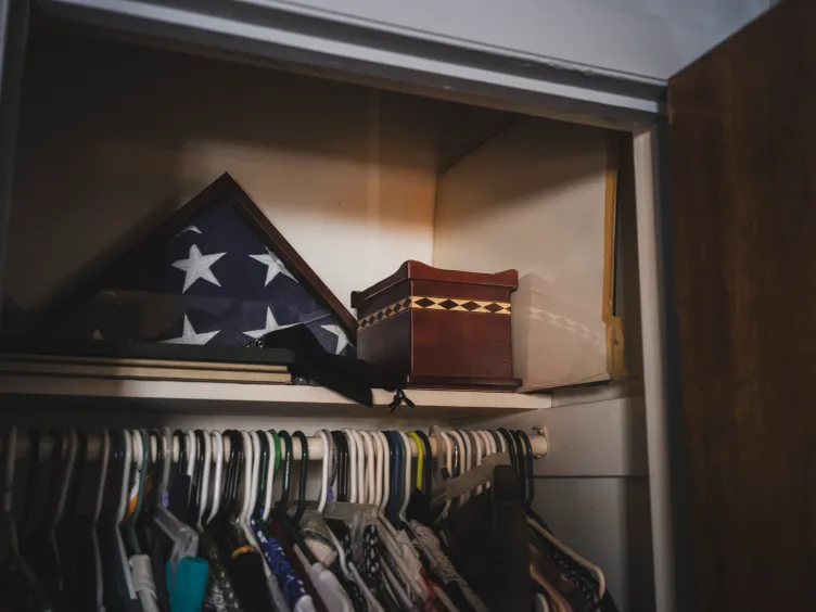 A small wooden box sits on a shelf inside a closet, next to an American flag and above clothing on hangers.