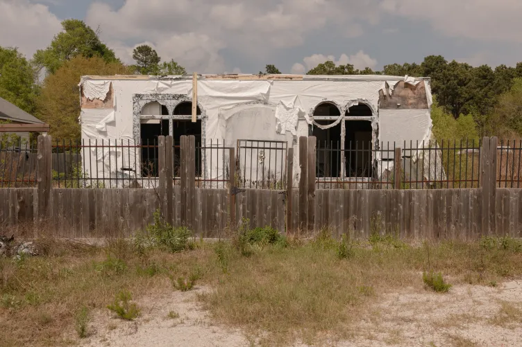 An unfinished house, draped with white plastic construction material, sits behind a deteriorated wooden fence.