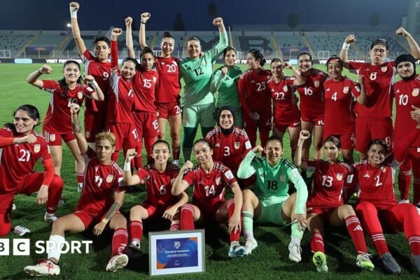 Players from the Afghan Women United team, half standing and half seated, pose for a post-match photo, smiling and with fists raised