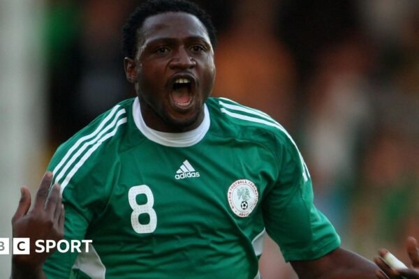Michael Eneramo shown from waist up, wearing a green Nigeria shirt with white trim, the number eight on the right of his chest and Nigeria crest on the left, holds his hands up and is seen with his mouth open in celebration after scoring a goal