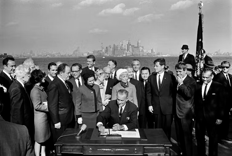 A man in a dark suit sits at a table, looking down at papers as several others around him look on.
