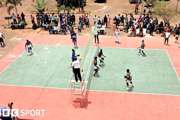 An elevated view gives a full picture of players competing in an outdoor Abula match on a mint green oblong court surrounded by a terracotta border, using the sport’s distinctive rectangular bats across a central net. A referee stands elevated at the centre to oversee proceedings across the net, while other officials and linesmen holding flags take their positions around the court. Spectators, both seated and standing on the far side of the court, watch intently from the sidelines, creating a lively atmosphere against the backdrop of trees, vehicles, and open space, while a man in a blue cap stands on a raised podium and films the game from the near side