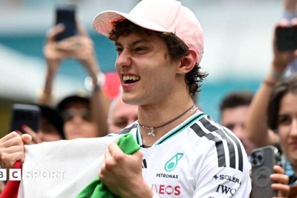 Kimi Antonelli celebrates his race win in Miami while holding the Italian flag. A crowd are gathered around him and taking photographs