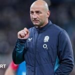 Steve Borthwick, wearing a nany blue zip-up England jacket, looks into the distance, his right hand raised to shoulder level