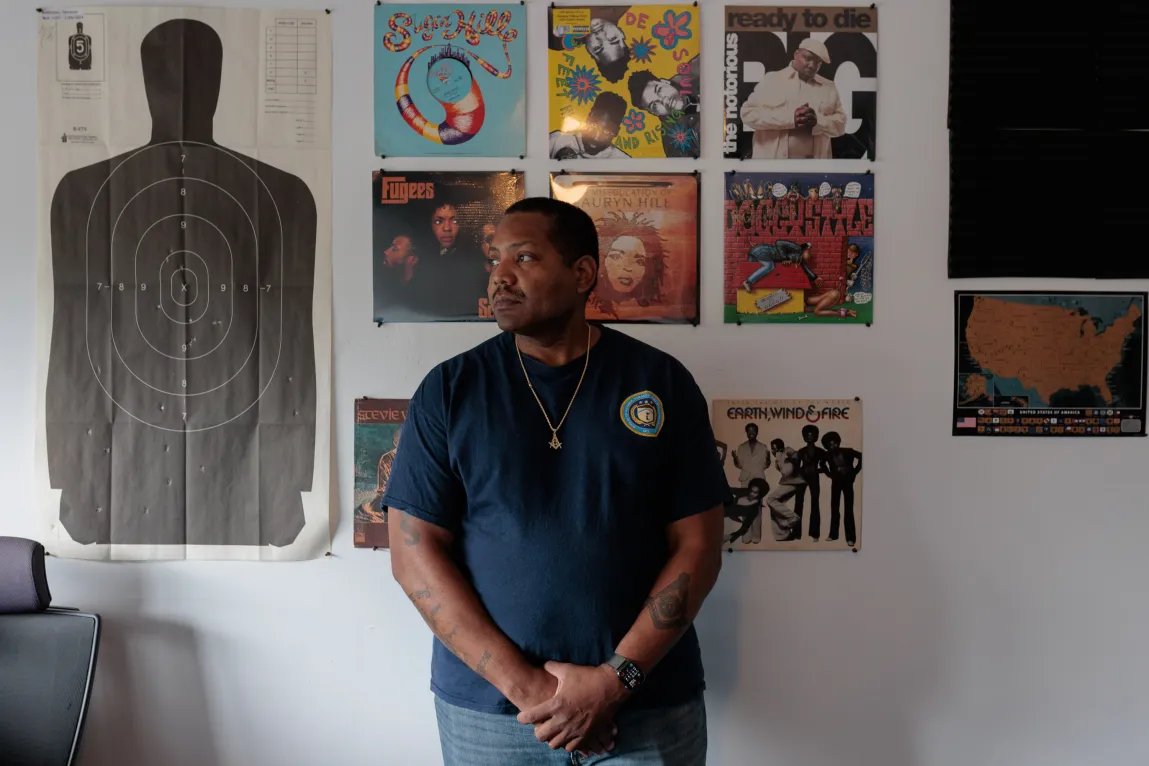 A man poses in front of a wall covered in album covers for vinyl records. To his left there is a paper poster of a silhouette made for shooting range practice.