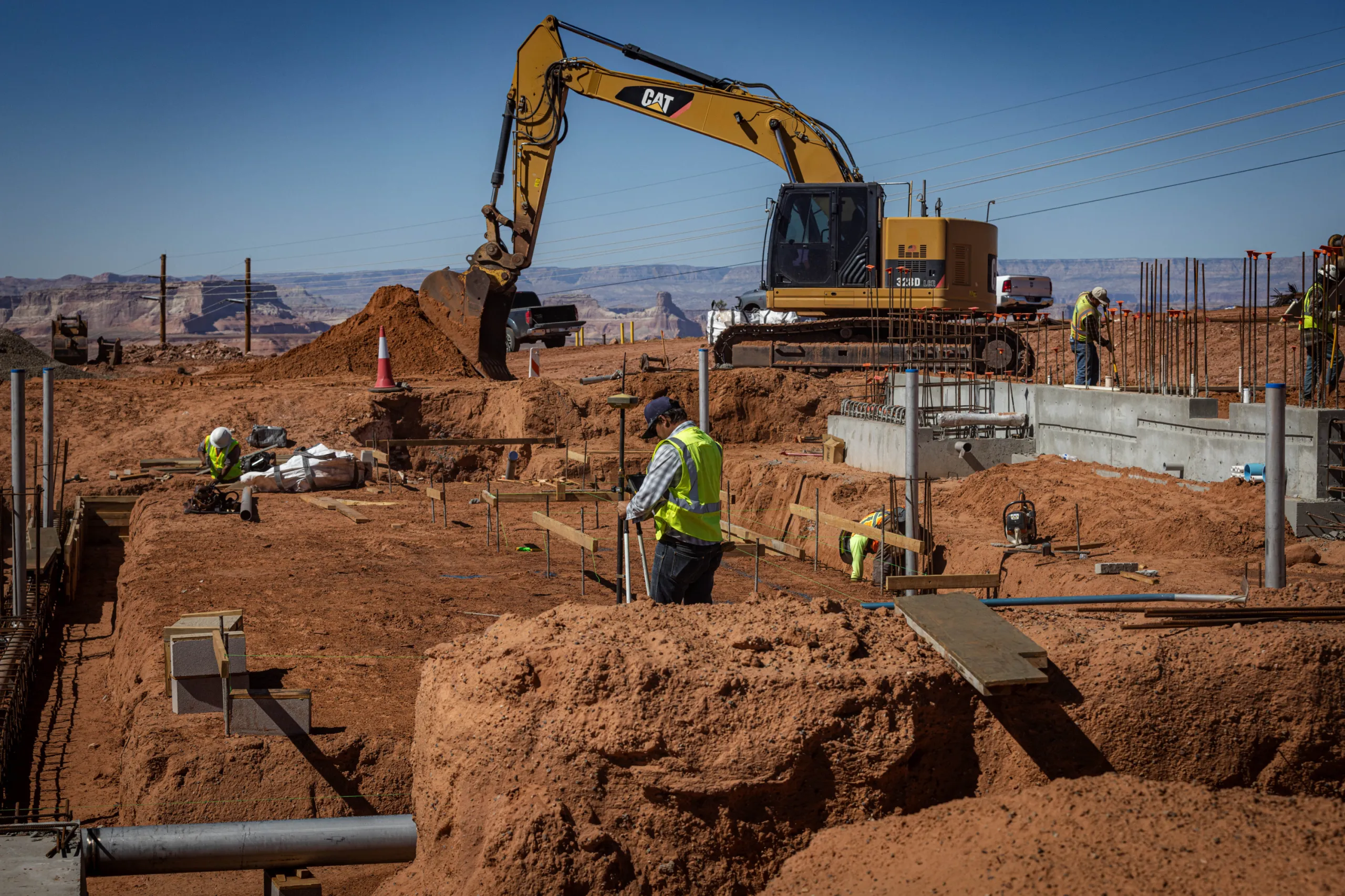 Construction workers in high-visibility vests work at a red-dirt excavation site alongside concrete foundations, while a large yellow excavator digs in the background.