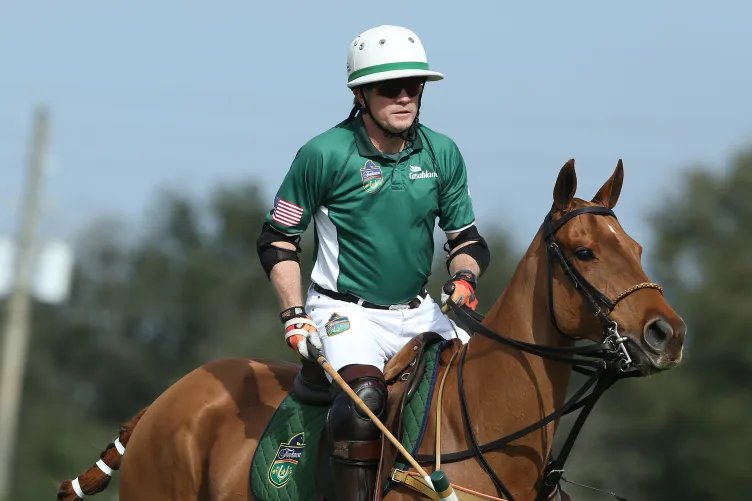 A man in a green jersey and helmet and holding a polo stick sits on a horse.