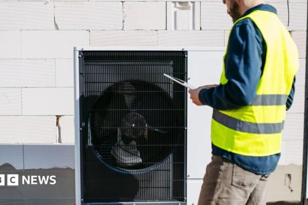 A black heat pump against a white breezeblock wall, a man stands to the right in brown jeans, blue shirt and a hi vis jacket holding a clipboard. He has a brown beard and the top of his head is cropped in the image