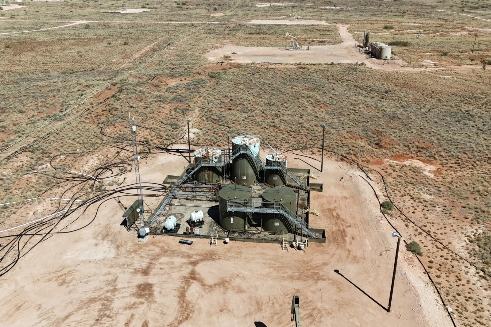 In a rural desert landscape there are large and rusty oil and gas storage tanks with pipes and tubes. Behind them are oil and gas pump jacks on cleared patches of land.