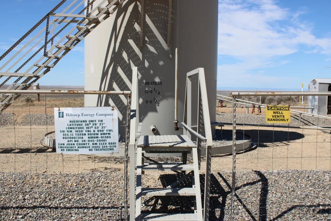 In a desert landscape there is a large, tan metal storage tank for oil and gas. It is surrounded by a fence. There are signs on the fence reading “Hilcorp Energy Company” and warning, “Caution: Well vents randomly.”