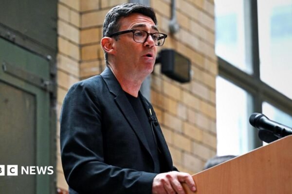 Andy Burnham, making a speech, wearing a dark T-shirt and grey jacket with a badge of a bee on the lapel.