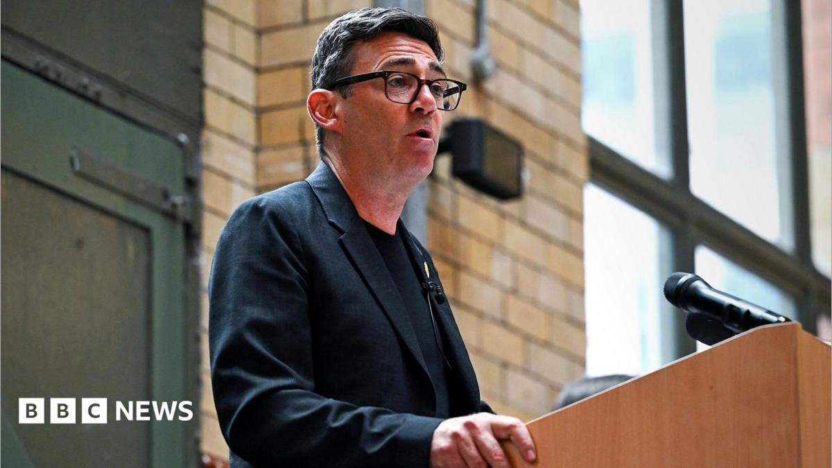 Andy Burnham, making a speech, wearing a dark T-shirt and grey jacket with a badge of a bee on the lapel.