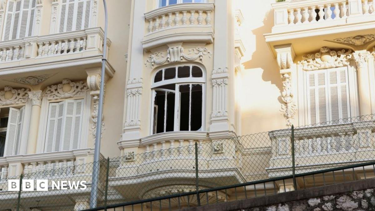 The damaged window of a light yellow residential building, which has balconies and ornate carvings.