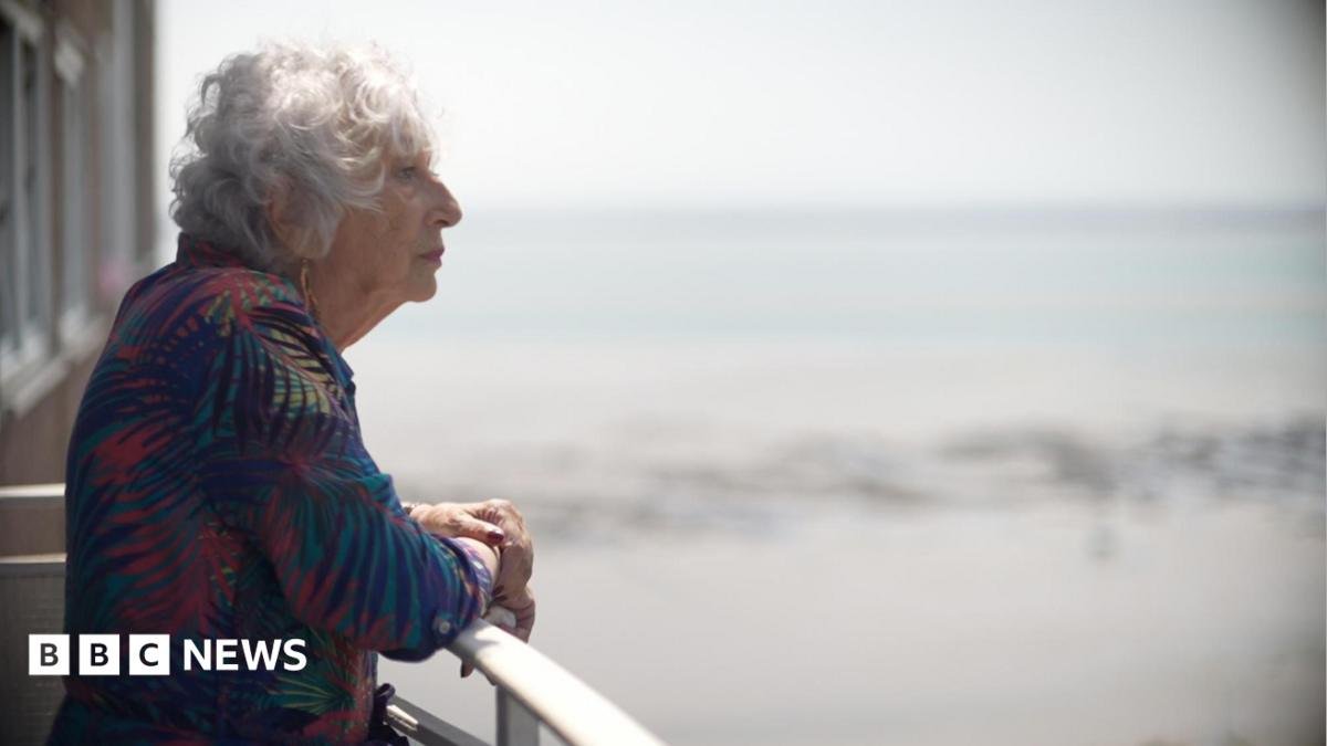 Carol Turansky standing on a balcony. She is wearing a leaf print dress and you can see her side profile. She has grey permed short hair and you can see the sea in the background. She is looking out to the horizon.