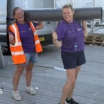 Two women, both holding their thumbs up, holding a carpet roll, both wearing shorts, and a purple T-shirt. The one on the left has on a high-vis jacket. A silver van is behind them and some wooden pallets.