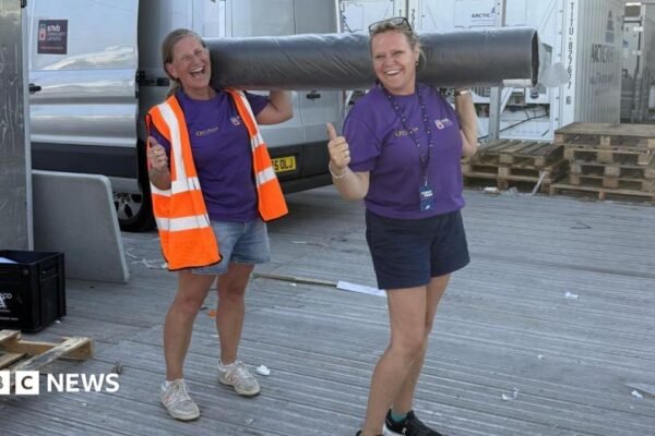 Two women, both holding their thumbs up, holding a carpet roll, both wearing shorts, and a purple T-shirt. The one on the left has on a high-vis jacket. A silver van is behind them and some wooden pallets.