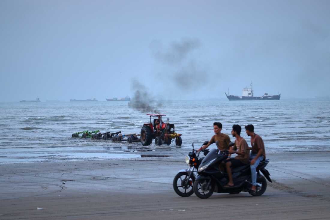 Young men ride motorcycles along the shore as a tractor pulls equipment from the water and commercial vessels appear anchored in the Strait of Hormuz off Bandar Abbas, Iran, Monday, July 27, 2026.