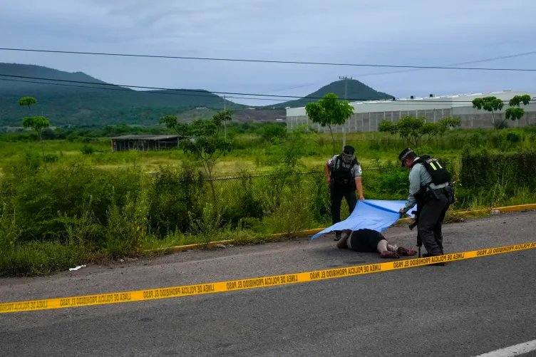 Two people in uniform drape a tarp over a dead body behind yellow police tape on the side of a road. The background is a lush green field with mountains in the distance.