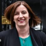 Phillipson wears a teal scoop-necked top with a black jacket and a small silver Celtic cross. She wears dark pink lipstick and is smiling. Her light brown hair is worn loose, straight and shoulder-length.