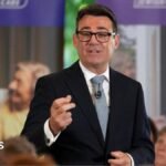 Andy Burnham is wearing a dark-coloured jacket, a white shirt, a tie and glasses. He is speaking to an audience who are not pictured.