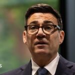A picture of Andy Burnham from the shoulders up. He is wearing a dark suit with a white shirt and dark tie, has thick-rimmed black glasses on, and has short, dark brown hair. The background behind him is blurred and he is looking forward with his mouth slightly open and his eyebrows raised.