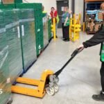 A FareShare worker moves palettes at the Middlesbrough hub. She is wearing black leggings and long-sleeved top, as well as a green vest. Her hair is straight and ginger.
