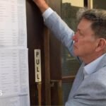 Ian Davidson posting the statement of persons nominated outside Clacton Town Hall. He is on the right, has short light brown hair and is wearing a light grey jacket over an open-neck shirt. His right arm is upraised as he puts the long list of typed names on to the brown wooden and glass door.