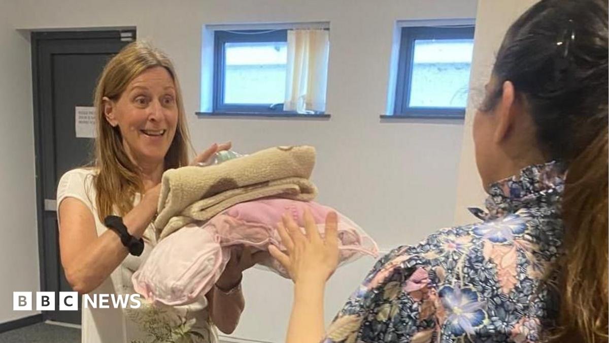 A woman holding a number of items that look like blankets, with a woman handing them to her. They both have long hair.