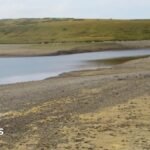 Llyn Aled Isaf in Conwy. The banks of the lake are dried up, the nearest bank is sandy with pebbles on, the bank in the distance is green, with a hill rising up behind it.