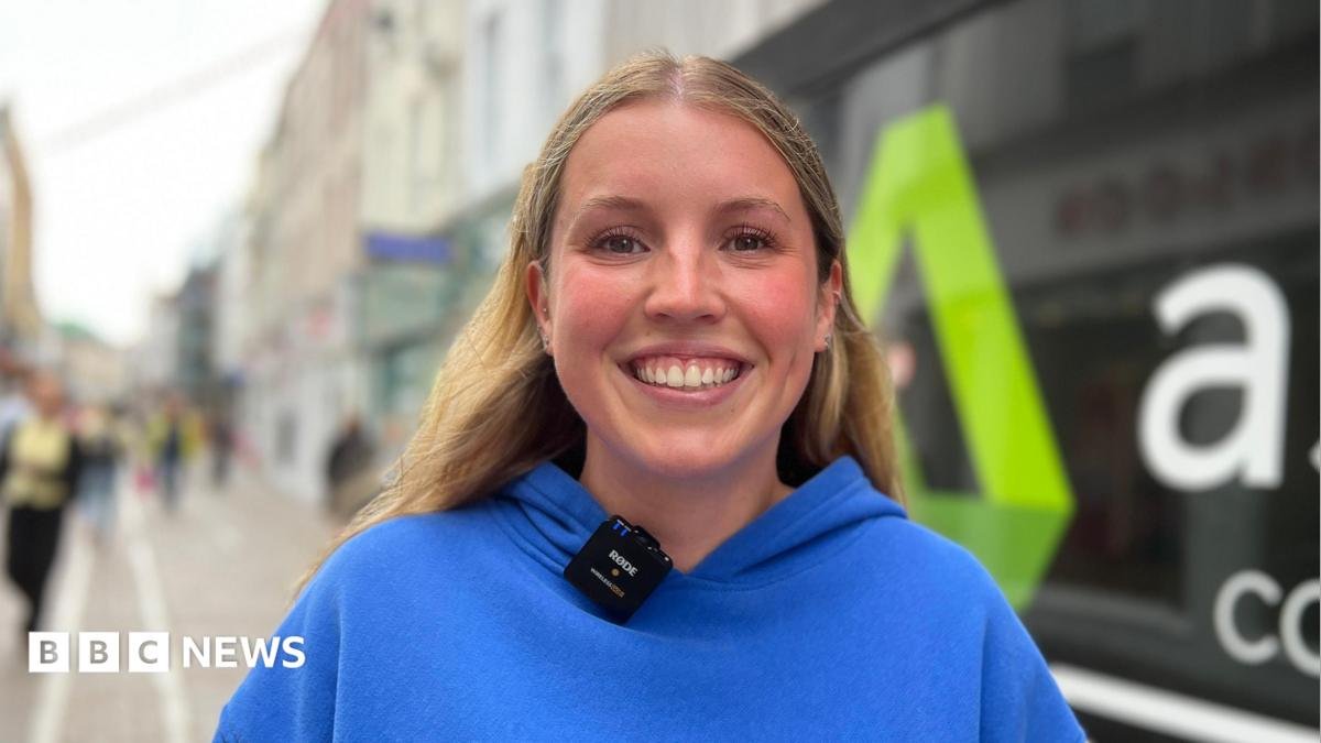 Eilidh is wearing a bright blue hoodie with the words “WHITE FOX” stands on a pedestrian shopping street. A small wireless microphone is clipped to the hoodie. Behind her, a shopfront window displays large white lettering and a green logo, while other pedestrians and buildings are visible in the softly blurred background.