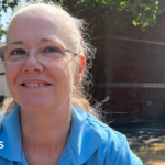 A woman with long blonde hair tied back is sitting on a wooden bench. She is wearing a blue polo shirt. She wears glasses.