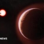 An artist's impression of exoplanet LHS 1140b, a large reddish-brown rocky planet filling the foreground, edged with a faint blue-white atmospheric glow. In the dark background, a small red star glows brightly, with a second, closer planet shown as a tiny black silhouette crossing in front of it