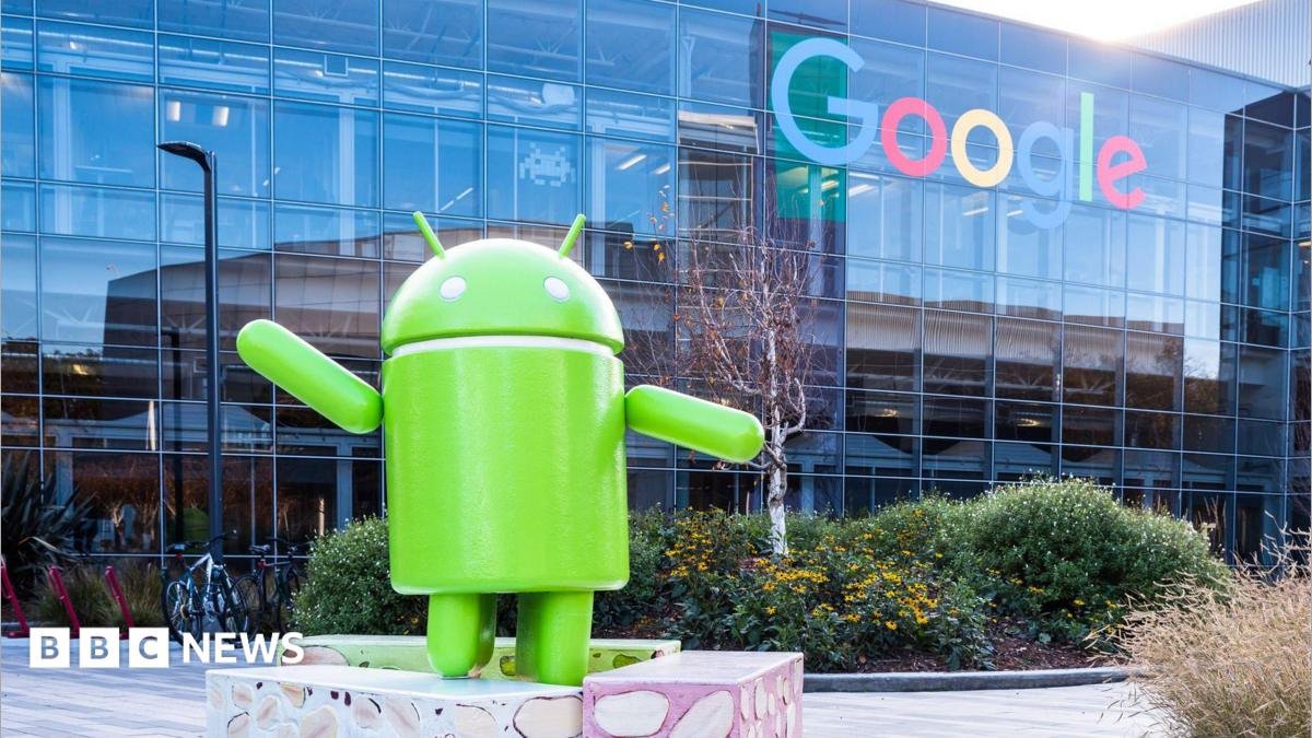 The green Android logo as a sculpture placed outside a glass Google office building, which has the Google logo on it.
