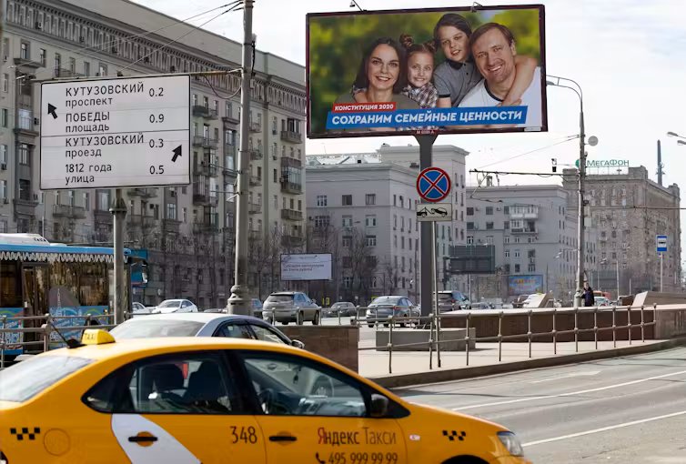 A yellow taxi drives past a billboard with a photo of a smiling woman, man and two children.