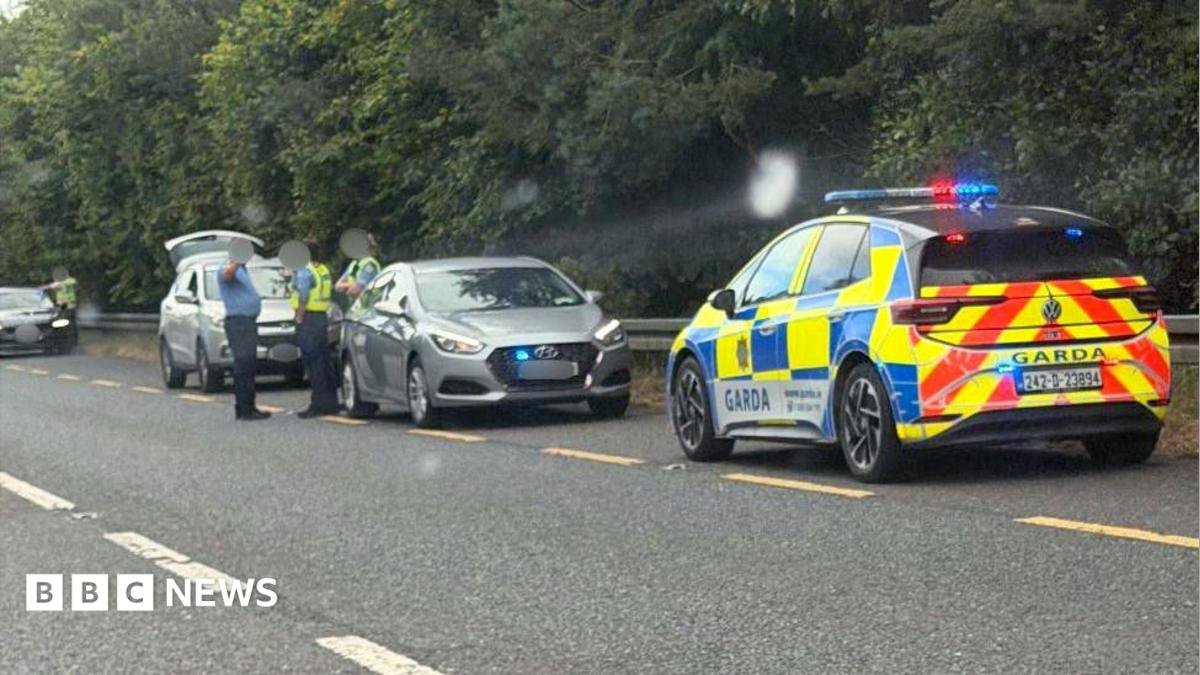 Garda officers and garda cars surround a small car on the roadside of the N2  near Carrickmacross. The officers are all wearing blue uniforms, but two are wearing yellow high-viz vests over their shirts.  The boot of a hatchback vehicle behind them is open.