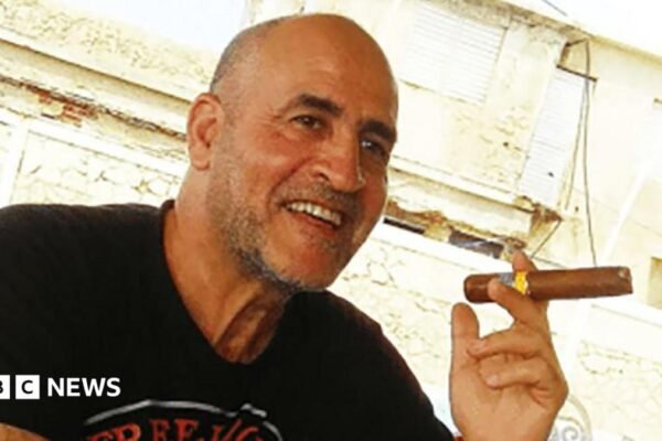 Daniel Siad, a man with a shaved head and stubble, wearing a black T-shirt, sits outside in what appears to be a sunny street, smiling with a large cigar raised in his hand.