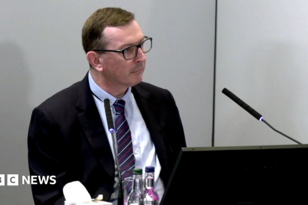 Brian O'Donnell sits behind a desk at the Lampard Inquiry at Arundel House in London. He is wearing a navy and burgundy tie and a black suit jacket, and a black laptop sits in front of him at the table. There are two bottles of water on the table and a microphone in front of him with a box of tissues.