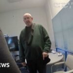 Murdoch wearing dark green jumper stands in front of camera while questioned by two officers in front of him