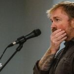 Graham Platner seen on stage at a microphone. his hand is over his mouth