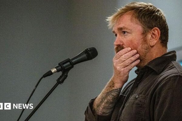 Graham Platner seen on stage at a microphone. his hand is over his mouth