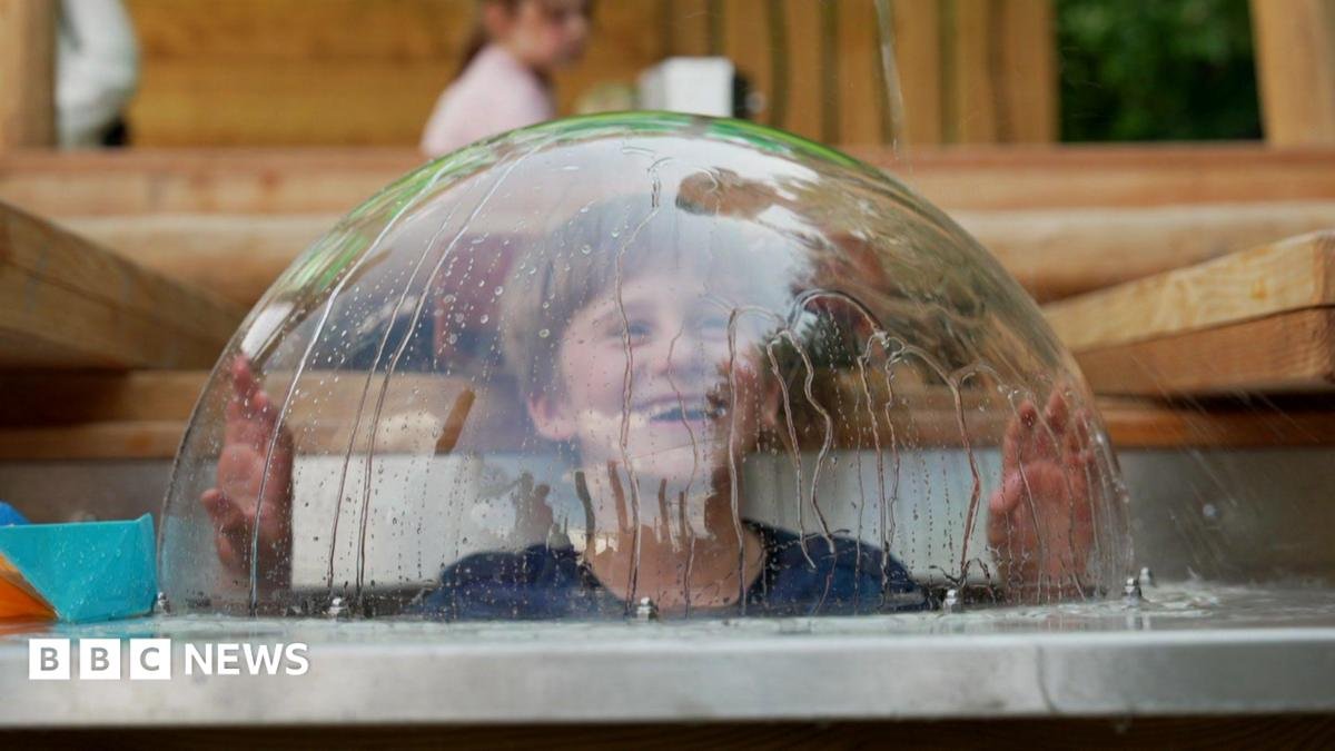 A boy pokes his head up in a glass lid, which has water dripping down the outside.