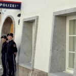 Two police officers stand outside a white-washed building with a police sign in German on the wall
