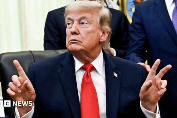 US President Donald Trump pointing fingers in the Oval Office of the White House, with an American flag and suited men behind him.