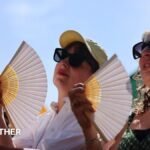 Two women in sunglasses fan themselves, with blue sky and sunshine overhead