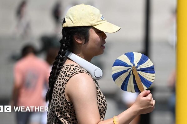 A woman cools herself with a handheld fan whilst walking in the street.