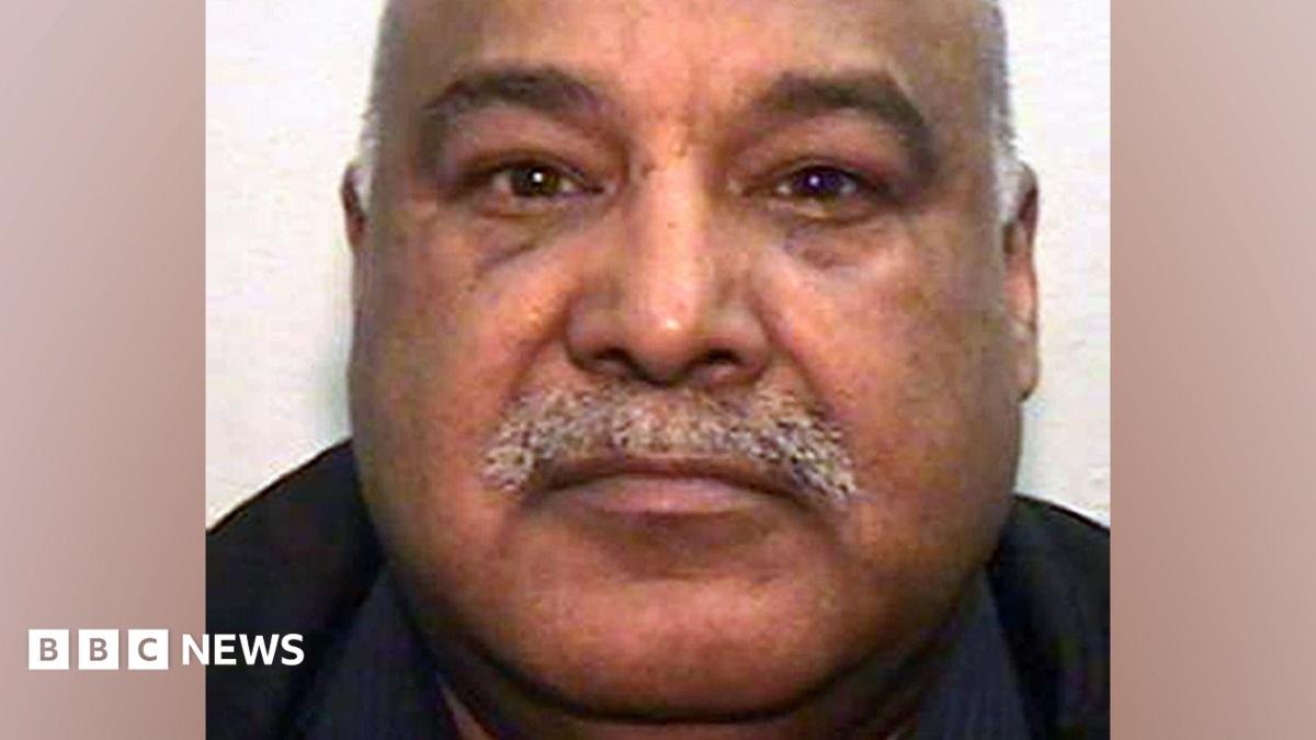 A mugshot of an Asian man in his 60s, with a grey moustache and bald head. He is looking into the camera.