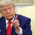 President Donald Trump pointing his finger. He has a serious look on his face and is wearing a blue suit with a white shirt and red tie.