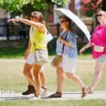 Four people strolling in the sunshine wearing shorts, one person is carrying an umbrella.